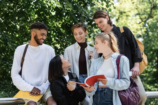 Asiatischer Student mit Imbissgetränk im Gespräch mit Freunden mit Notizbüchern im Park — Stockfoto