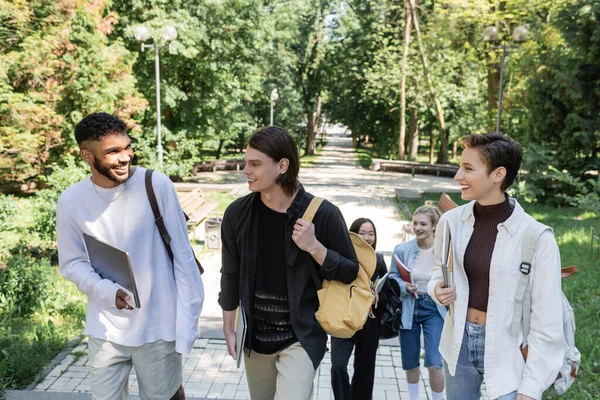 Étudiants interraciaux avec appareils et sacs à dos marchant dans le parc — Photo de stock