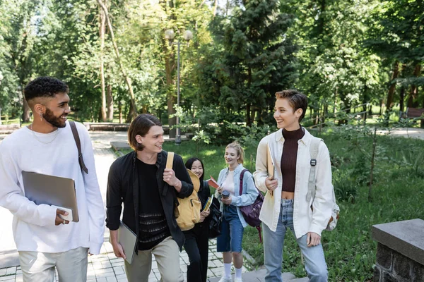 Estudantes multiétnicos positivos com dispositivos conversando enquanto caminham no parque — Fotografia de Stock