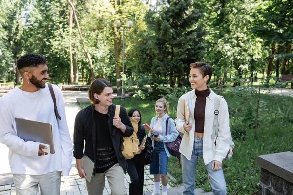 Étudiants multiethniques positifs avec des appareils parlant tout en marchant dans le parc — Photo de stock