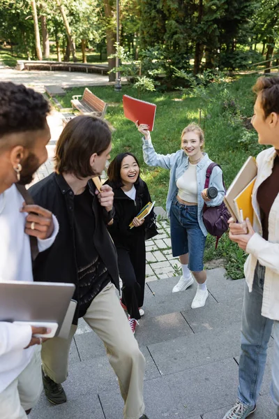 Interracial students with devices looking at cheerful friends in park — Stock Photo