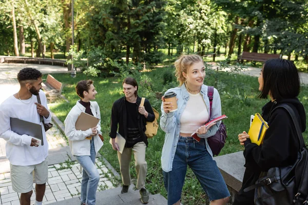 Estudantes inter-raciais positivos com dispositivos e cadernos falando no parque — Fotografia de Stock