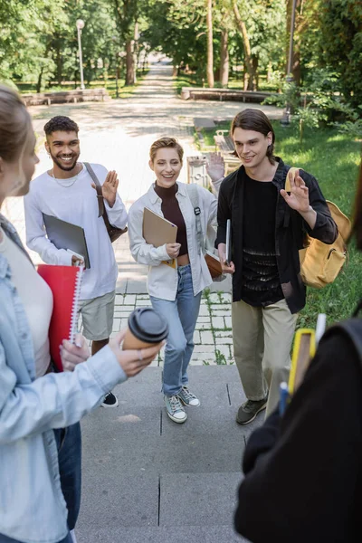 Estudiantes interracial positivos con gadgets saludando a amigos en el parque - foto de stock