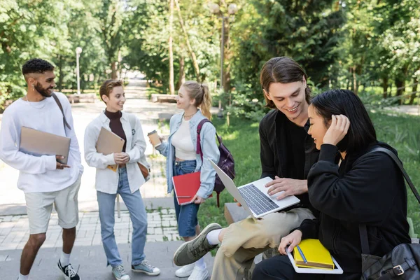 Smiling student holding laptop near asian friend in summer park — Stock Photo