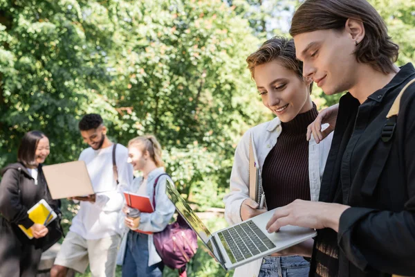 Lächelnde Studenten mit Laptop in der Nähe von verschwommenen interrassischen Freunden im Park — Stockfoto