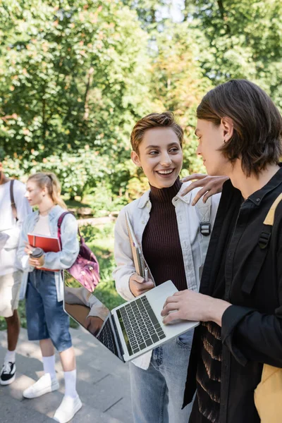 Positive students with laptop talking near blurred multiethnic friends in park — Foto stock