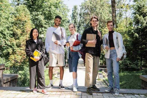 Estudantes multiculturais sorridentes com cadernos e laptops olhando para a câmera no parque — Fotografia de Stock