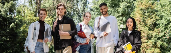 Alegre estudiantes interracial con aparatos y cuadernos mirando a la cámara en el parque de verano, pancarta - foto de stock