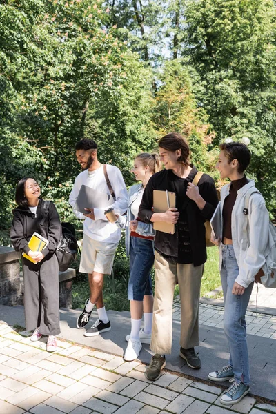 Smiling interracial students looking at cheerful asian friend with notebooks in park — Stock Photo