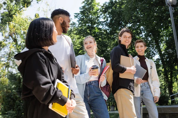 Низький кут зору багатоетнічних студентів з блокнотами, що розмовляють в літньому парку — стокове фото