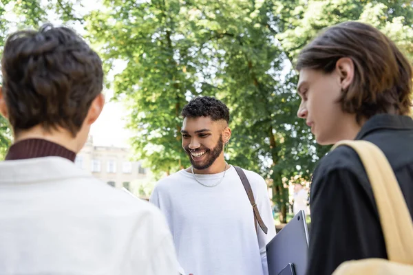 Positive african american student holding laptop near blurred friends in park — Stock Photo