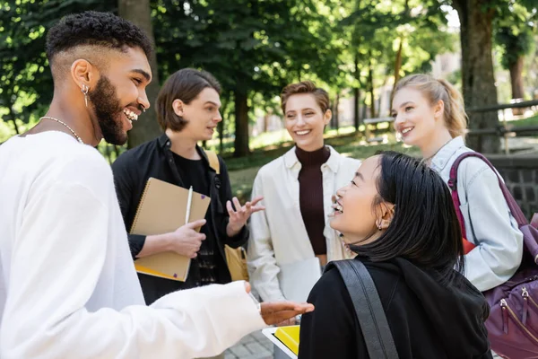 Lächelnde gemischtrassige Studenten unterhalten sich in der Nähe verschwommener Freunde mit Rucksäcken im Sommerpark — Stockfoto