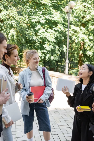 Smiling students with notebooks looking at asian friend with digital tablet in park — Stockfoto