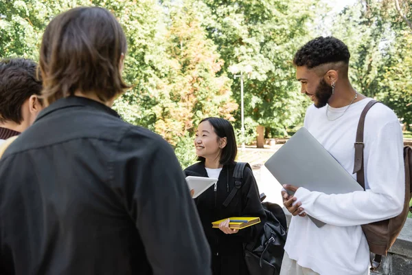 Smiling african american student holding laptop and looking at asian friend with digital tablet in park — Stock Photo