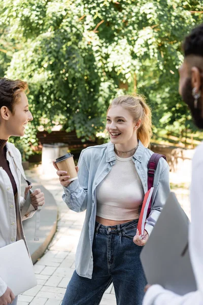 Estudiante sonriente sosteniendo café para ir y cuaderno cerca de amigos interracial en el parque - foto de stock