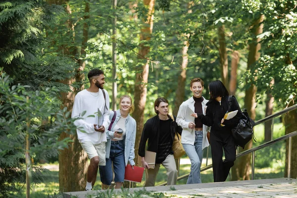 Positive interracial students with notebooks walking on stairs in park — Stockfoto