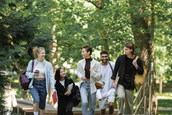 Fröhliche Schüler mit Rucksäcken spazieren in der Nähe multiethnischer Freunde im Park — Stockfoto