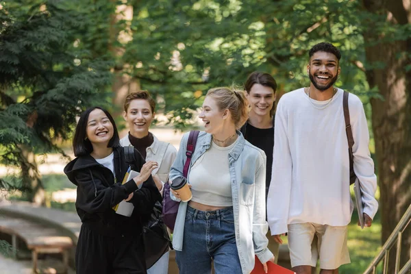 Smiling asian student walking near multicultural friends in summer park - foto de stock