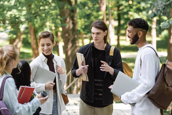 Positive interracial students with laptops talking to blurred friends in park - foto de stock
