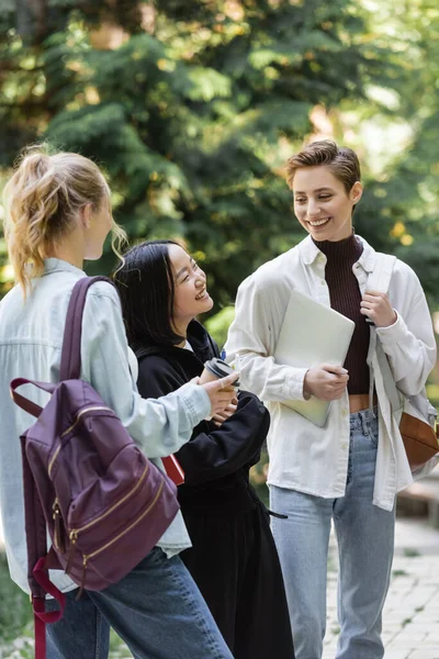 Smiling asian student standing near friends with backpacks in park — Stock Photo