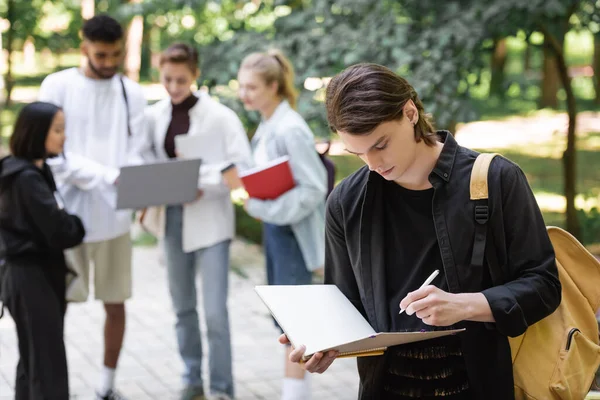 Estudiante sosteniendo cuaderno cerca borrosa interracial amigos en parque - foto de stock