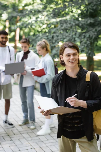 Smiling student holding notebook near blurred interracial friends in park - foto de stock