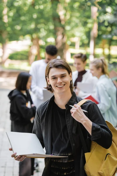 Studente sorridente con taccuino e zaino guardando la fotocamera vicino agli amici sfocati nel parco — Foto stock