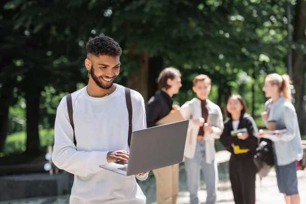 Estudante afro-americano sorrindo usando laptop perto de amigos desfocados no parque — Fotografia de Stock