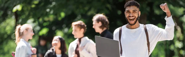 Excited african american student holding laptop near blurred friends in summer park, banner — Stock Photo
