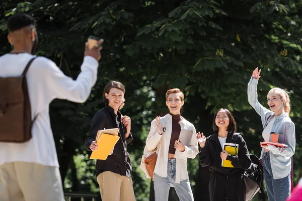 Positive interracial students waving hands near blurred african american friend in park — Stock Photo