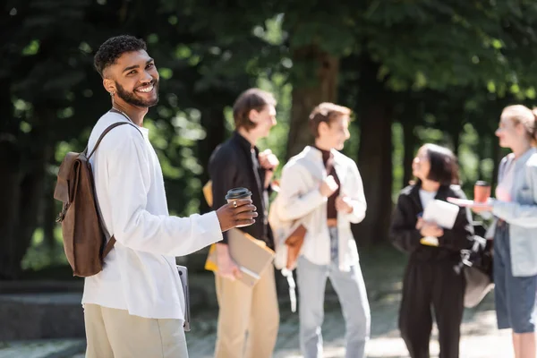 Sorrindo estudante afro-americano segurando café para ir perto de amigos embaçados no parque — Fotografia de Stock