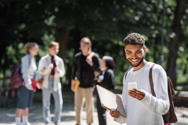 Étudiant afro-américain souriant utilisant un smartphone et tenant un carnet dans le parc — Photo de stock