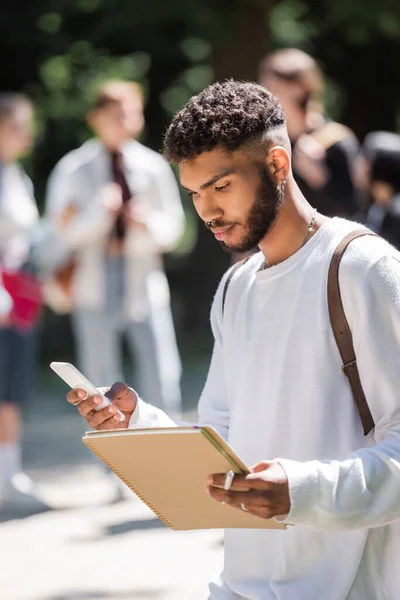 African american student holding smartphone and notebook in park — Photo de stock