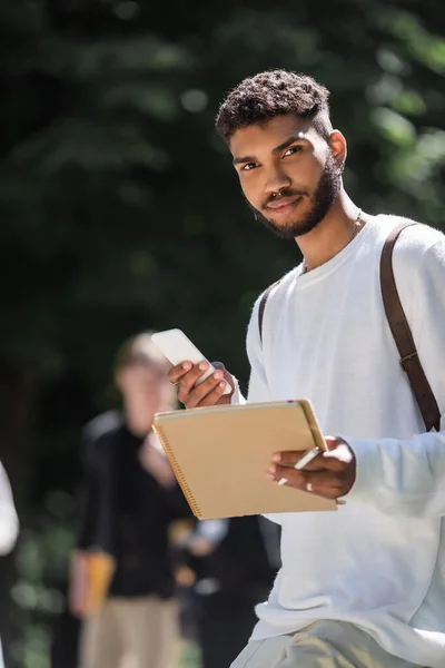 Jeune étudiant afro-américain tenant smartphone et ordinateur portable dans le parc — Photo de stock