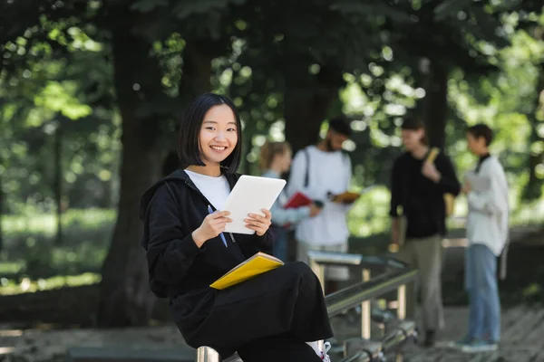 Fröhlicher asiatischer Student mit digitalem Tablet und Notizbuch blickt im Park auf Kamera — Stockfoto