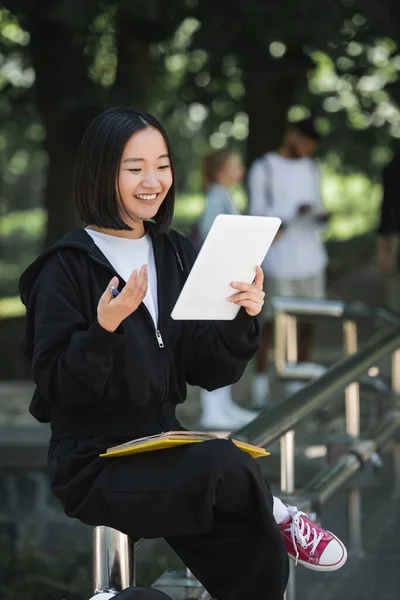Sonriente asiático estudiante teniendo vídeo llamada en digital tablet en parque - foto de stock
