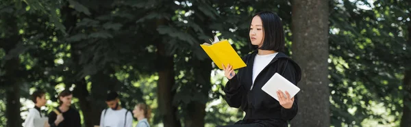 Азійський студент тримає в парку цифрову табличку і книжку для читання. — стокове фото