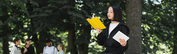 Asian student holding digital tablet and reading book in park, banner — Stock Photo