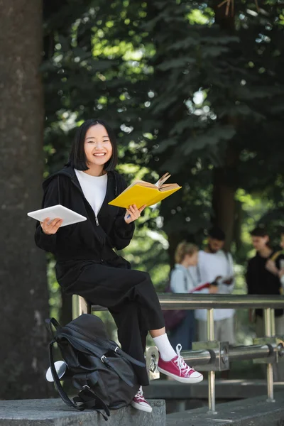 Cheerful asian student with book and digital tablet looking at camera near backpack in park - foto de stock