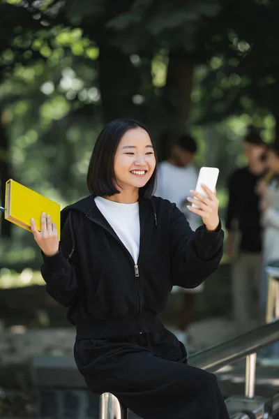 Smiling asian student holding book and taking selfie on smartphone in park - foto de stock