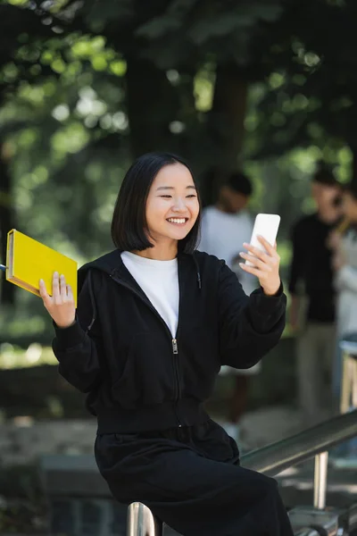 Smiling asian student holding book and taking selfie on smartphone in park — Stock Photo