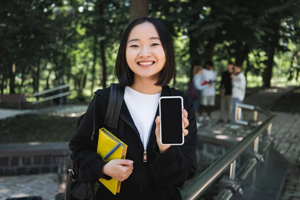 Cheerful asian student with book showing smartphone in park - foto de stock