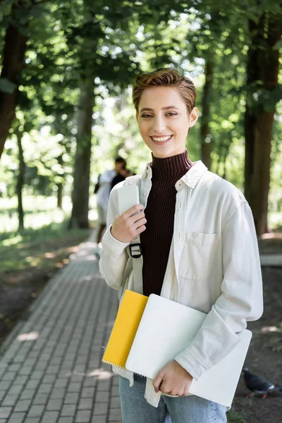 Estudante positivo segurando notebook e dispositivos no parque de verão — Fotografia de Stock