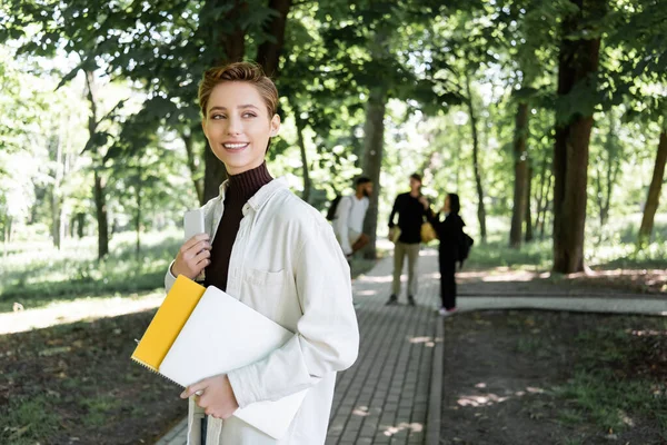 Cheerful student holding gadgets and notebook in summer park — Fotografia de Stock