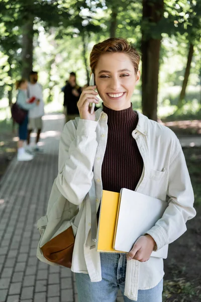 Positive student with laptop and notebook talking on smartphone in park - foto de stock