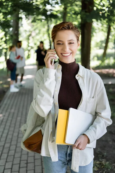 Étudiant positif avec ordinateur portable et ordinateur portable parlant sur smartphone dans le parc — Photo de stock