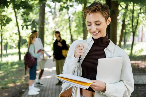 Fröhlicher Student mit Laptop schaut im Sommerpark auf Notizbuch — Stockfoto