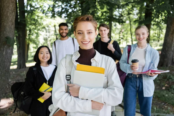 Studente sorridente con computer portatile e notebook guardando la fotocamera vicino a sfocati amici interrazziale nel parco — Foto stock