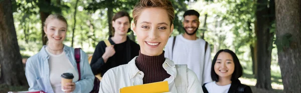 Позитивный студент с ноутбуком, смотрящий в камеру рядом с мультикультурными друзьями в летнем парке, баннер — стоковое фото