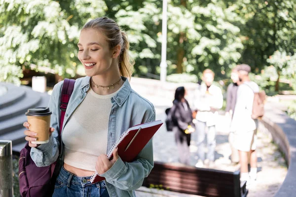 Estudante sorrindo segurando café para ir e notebook no parque — Fotografia de Stock